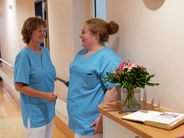 Zwei Pflegekräfte der Palliativpflege unterhalten sich in blauer Funktionskleidung auf einem hellen Stationsflur neben einem mit Blumen geschmückten Stationstisch in der Klinik für Innere Medizin mit Schwerpunkt Palliativmedizin im Albertinen Krankenhaus in Hamburg. Zwei Pflegekräfte der Palliativpflege unterhalten sich in blauer Funktionskleidung auf einem hellen Stationsflur neben einem mit Blumen geschmückten Stationstisch in der Klinik für Innere Medizin mit Schwerpunkt Palliativmedizin im Albertinen Krankenhaus in Hamburg.