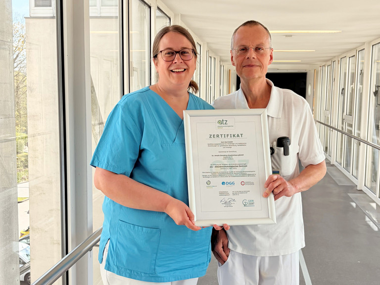 Dr. Saskia Otte, Chefärztin der Geriatrie und Physikalischen Medizin, und Dr. Olaf Pingen, Chefarzt der Orthopädie und Unfallchirurgie, stehen im Amalie Sieveking Krankenhaus in Hamburg-Volksdorf in einem hellen Durchgang und halten gemeinsam das Zertifikat für die erfolgreiche Zertifizierung des Krankenhauses als Alterstraumatologisches Zentrum in die Kamera. Dr. Saskia Otte, Chefärztin der Geriatrie und Physikalischen Medizin, und Dr. Olaf Pingen, Chefarzt der Orthopädie und Unfallchirurgie, stehen im Amalie Sieveking Krankenhaus in Hamburg-Volksdorf in einem hellen Durchgang und halten gemeinsam das Zertifikat für die erfolgreiche Zertifizierung des Krankenhauses als Alterstraumatologisches Zentrum in die Kamera.